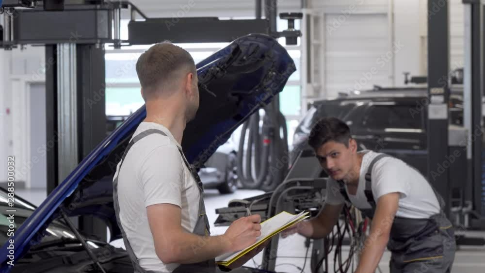 Two mechanics discuss repairing broken automobile at the garage. Male technician making notes on his clipboard, talking to his assistant examining motor of an auto. Workshop concept Two mechanics discuss repairing broken automobile at the garage. Male technician making notes on his clipboard, talking to his assistant examining motor of an auto. Workshop concept