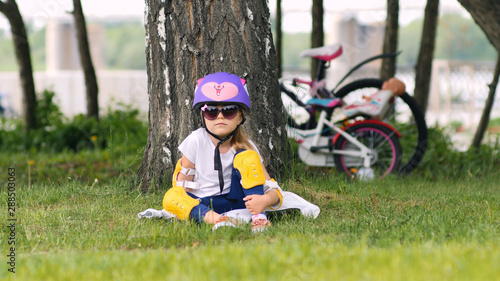 Cute Little Caucasian Girl Wearing Bicycle Helmet Is Sitting Under The Tree In The Park After Bike Ride. Bicycle At The Background In City Park