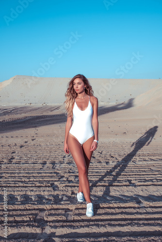A girl in a white bathing suit, walks in the summer on white sand, against the backdrop of the hills by the sea, a weekend walk by the lake. Long hair tanned figure.