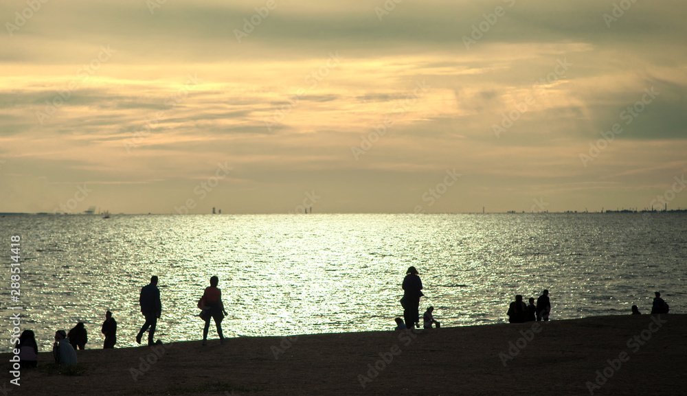 Fototapeta premium Silhouettes of people walking along the shore of the sea bay against the sunset sky with clouds.