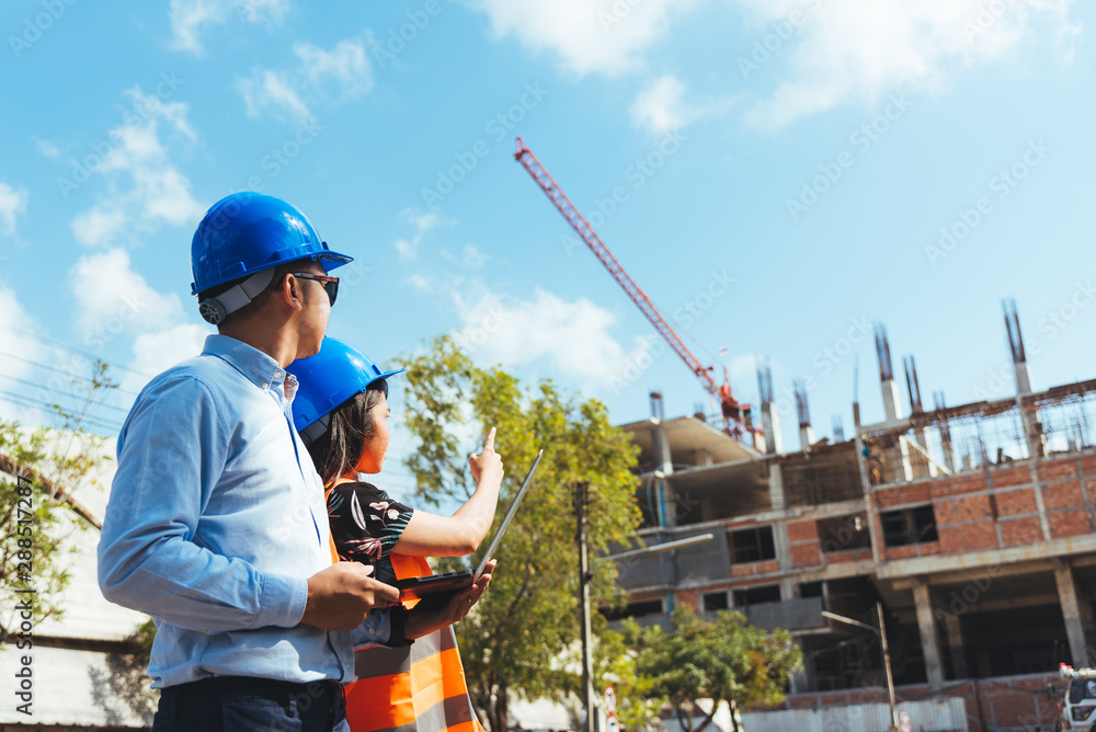 Asian man and woman engineer with the blue safety helmet meeting at the construction site with blueprints and laptop. Asian man and woman engineer with the blue safety helmet meeting at the construction site with blueprints and laptop.