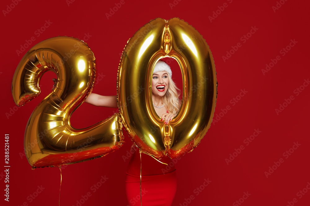 Portrait of young beautiful woman in fashion dress posing with gold 20 twenty number balloons isolated over red background. Christmas and new year Credit and deposit installments concept. Portrait of young beautiful woman in fashion dress posing with gold 20 twenty number balloons isolated over red background. Christmas and new year Credit and deposit installments concept.