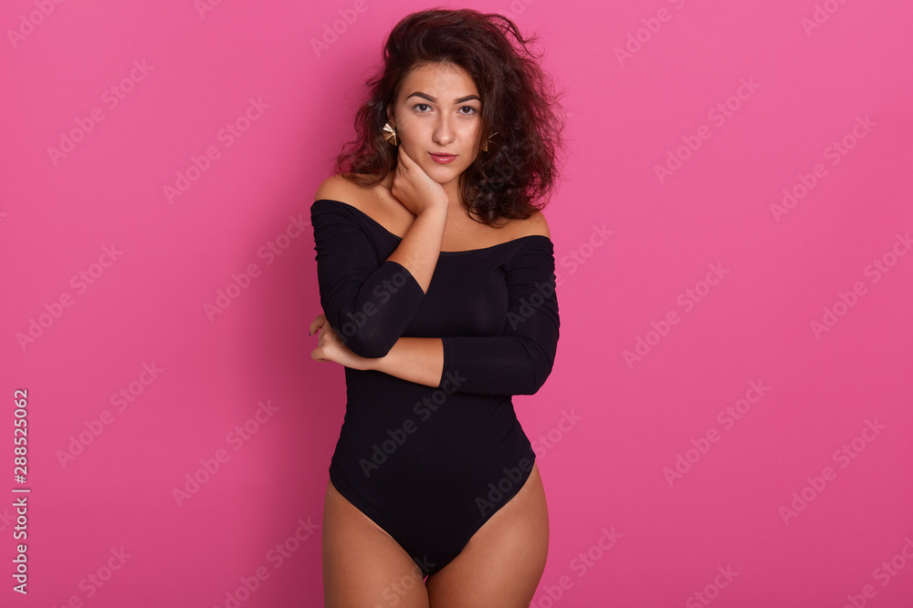 Obraz premium Brunette tanned female with hand touching her neck, having wavy hair. Portrait over rosy studio background, attractive girl wearing black combidress, looking directly atcamera, looks confident.
