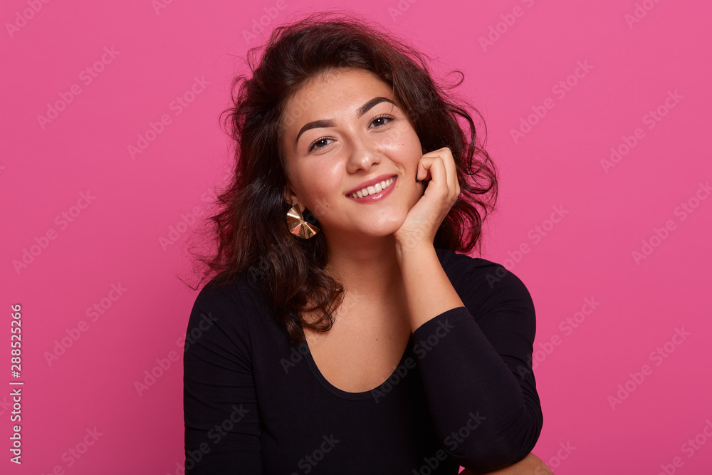 Indoor shot of beautiful girl smiling and looking directly atcamera, keeps hand under her chin. model posing isolated over rosy background, attractive lady has happy facial expression. People concept. Indoor shot of beautiful girl smiling and looking directly atcamera, keeps hand under her chin. model posing isolated over rosy background, attractive lady has happy facial expression. People concept.