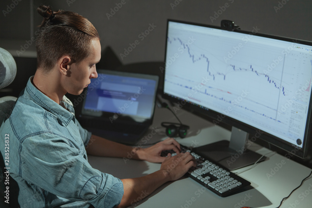 Young male programmer working late at night, copy space. Male stock trader using two computers, working in a dark office room