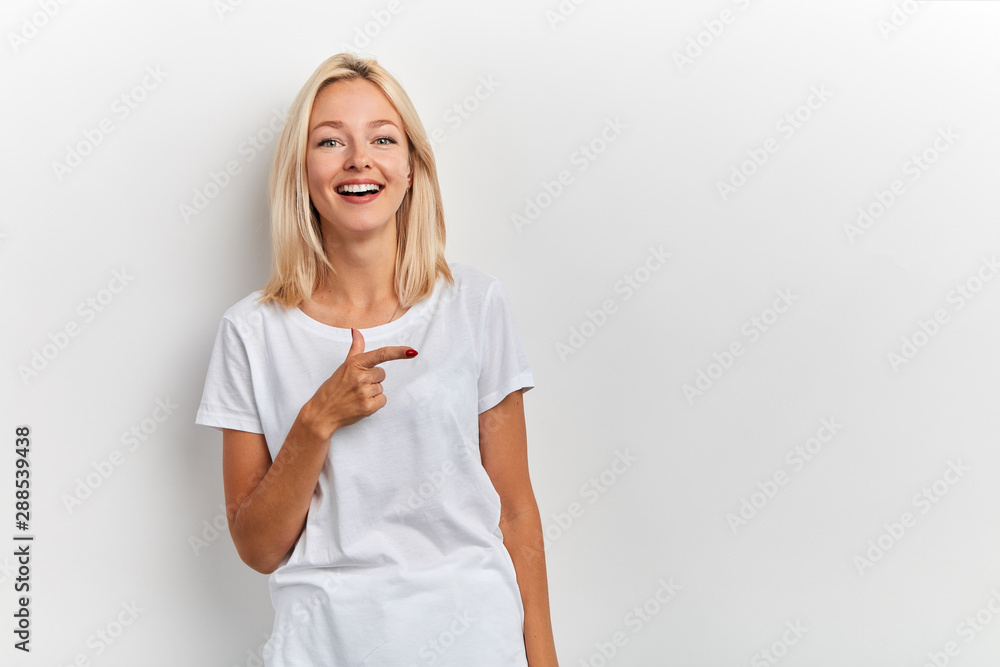 Happy charming young woman has joyful expression, points with finger aside, wears casual t shirt, isolated over white background with empty space. copy space. facial expression, direction, this way Happy charming young woman has joyful expression, points with finger aside, wears casual t shirt, isolated over white background with empty space. copy space. facial expression, direction, this way