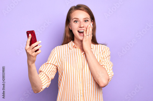 Front view of suprised funny young woman smiling broadly, holding red cell phone, cant believe in her success, happy and emotional, dressed in stylish shirt, standing at light purple wall.