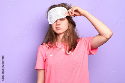 Front view of exhausted young woman raises slightly her eye mask, trying to open sleepy eyes, needs more rest after hard week, out of energy, hates morning. Sleep and wake up concept.