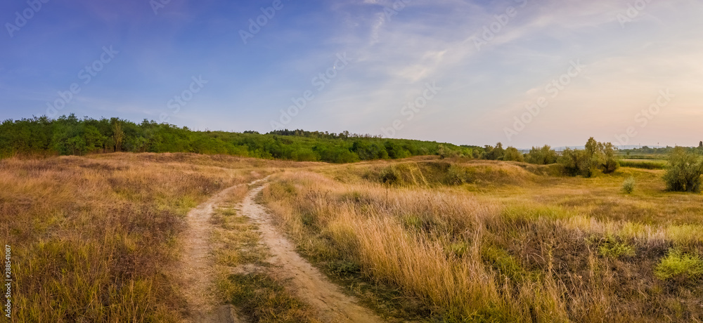 Idyllic autumn rural panorama with and a country track across a meadow with dry grass and hay. Beautiful evening scene, peaceful sunset light over the steppe vegetation.