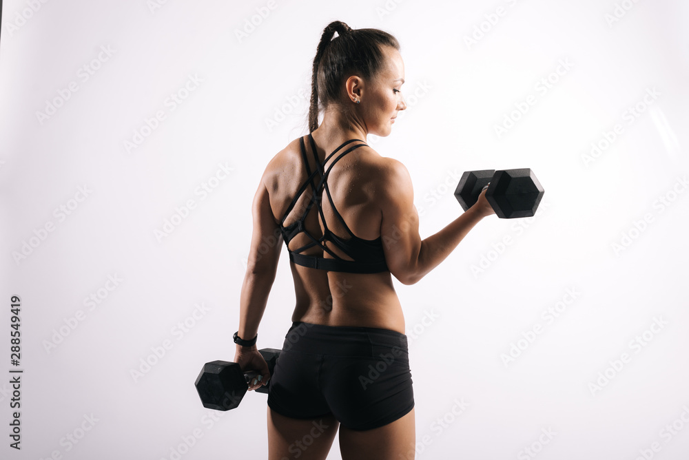 Back view portrait of fitness female with perfect muscular body in black sportswear. Working out with a heavy dumbbell on isolated white background Back view portrait of fitness female with perfect muscular body in black sportswear. Working out with a heavy dumbbell on isolated white background