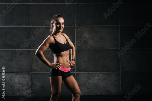 Young muscular woman bodybuilder in black sportswear is standing with hands on her belt, on black background. Sporty female looking at camera © dikushin