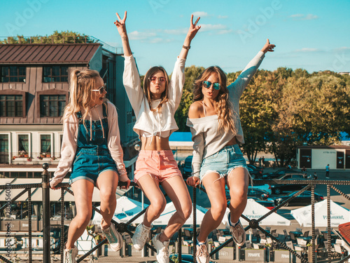 Portrait of three young beautiful smiling hipster girls in trendy summer clothes.Sexy carefree women sitting on handrail the street.Positive models having fun in sunglasses.They raising hands