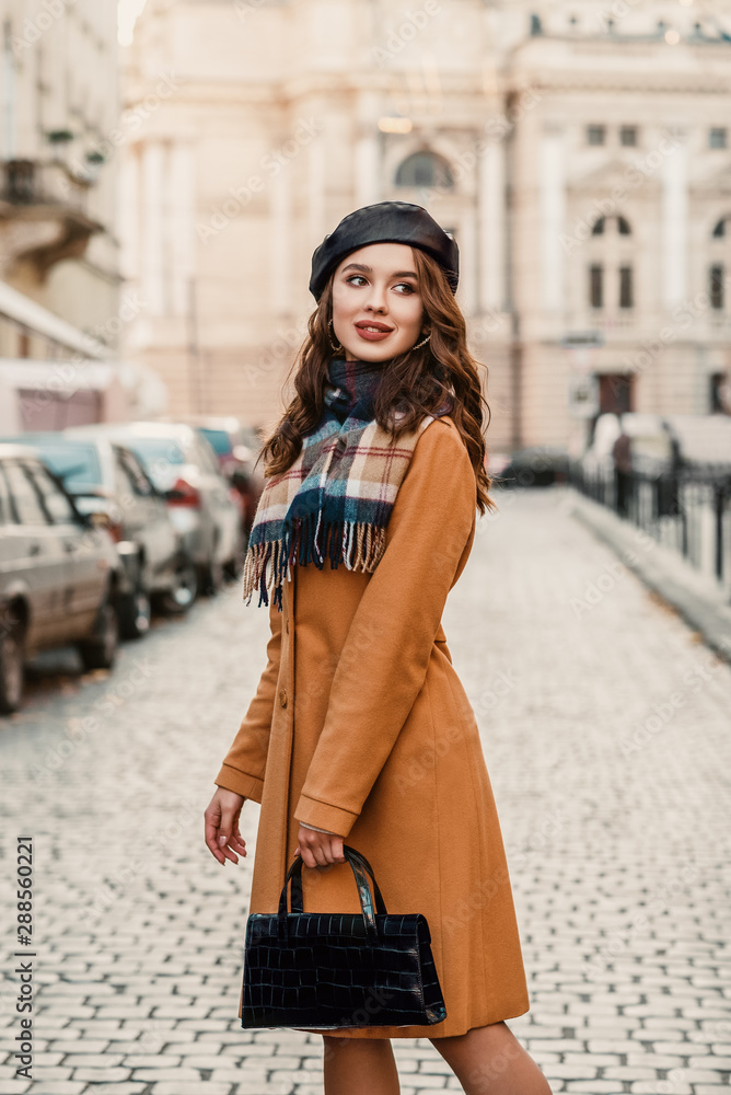 Outdoor autumn portrait of young happy smiling elegant fashionable lady wearing trendy beret, camel color coat, plaid scarf, holding faux croco leather baguette bag, walking in street of European city