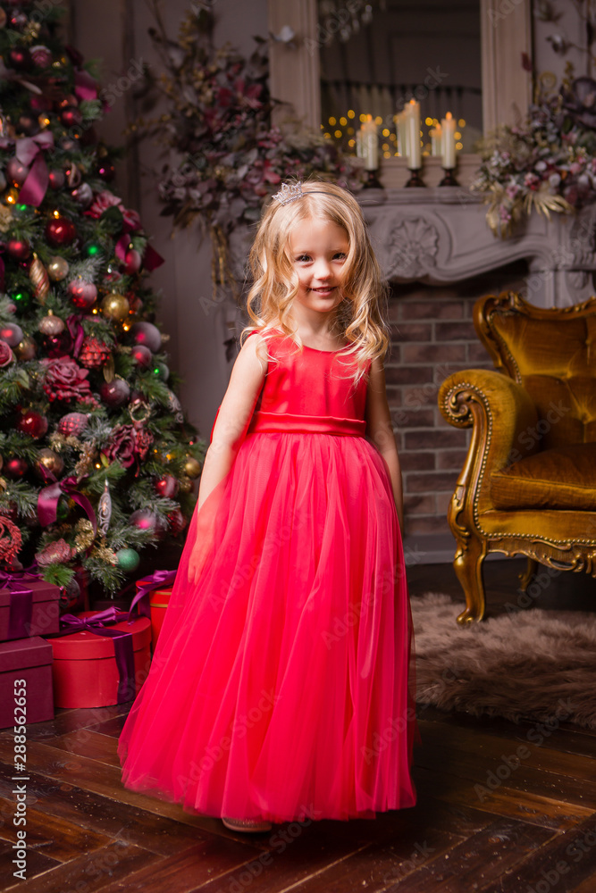 little blonde girl in evening red dress near the Christmas tree, child with Christmas gifts, the night before Christmas little blonde girl in evening red dress near the Christmas tree, child with Christmas gifts, the night before Christmas