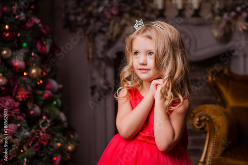 little blonde girl in evening red dress near the Christmas tree, child with Christmas gifts, the night before Christmas