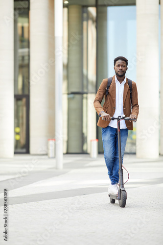 Full length portrait of contemporary African-American man riding electric scooter and looking at camera while commuting in city streets, copy space