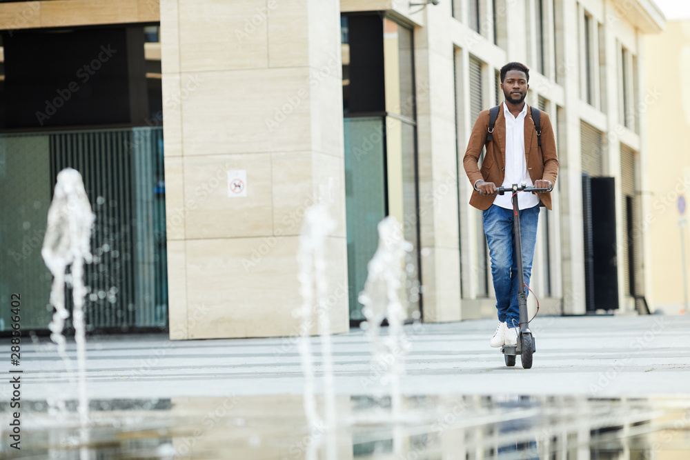 Front view portrait of contemporary African-American man riding electric scooter in urban setting outdoors, copy space Front view portrait of contemporary African-American man riding electric scooter in urban setting outdoors, copy space