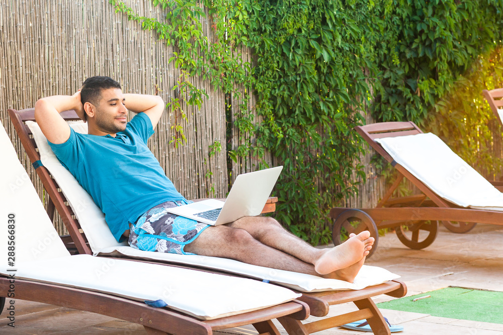 Portrait of pleasure bearded young adult freelancer man in blue t-shirt and shorts lying on cozy daybed with laptop on poolside and watching film online. Lifestyle concept, outdoor, summer vacation