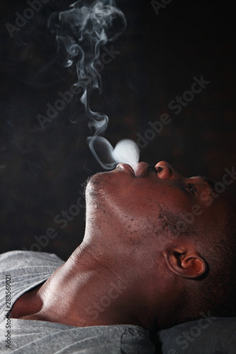 An African American man exhales smoke from a hookah. Portrait on a dark background in the hookah room interior.Not good habits.