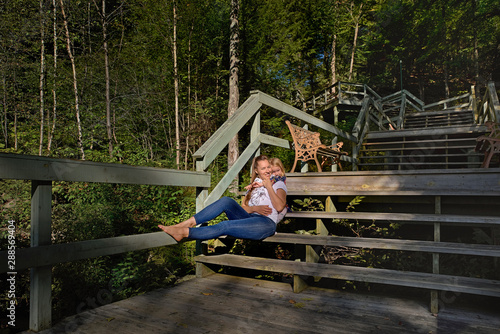 Mom and daughter are resting sitting on the steps on the trail in the autumn forest. Little girl hugs mom by the neck. Family walk and happy family.