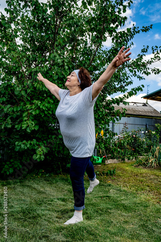Senior woman practice yoga on the lawn. She stands on one leg and throws up her hands to the side. She is happy and smiles. Old ages healthy lifestyle concept. Green tree on the background. Green lawn