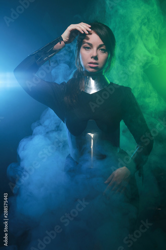 futuristic fashion model wearing black and silver clothes and standing in the colorful blue and green smoke