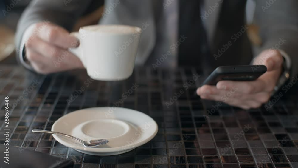Male businessman in suit use smartphone, business chatting while sitting at table in cafe. Cup of cappuccino coffee in front of camera. Coffee break, rest Male businessman in suit use smartphone, business chatting while sitting at table in cafe. Cup of cappuccino coffee in front of camera. Coffee break, rest