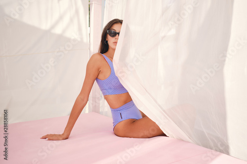 Gorgeous brunette girl in swimsuit and sunglasses thoughtfully covering half of face with curtains on beach bed