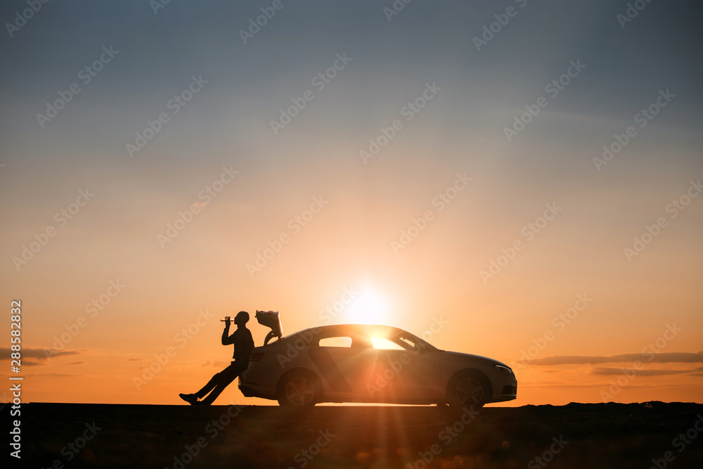 Silhouette of man driver relaxing after a ride, sitting on the trunk of his car and drinking water from a bottle, side view. Sunset time. 