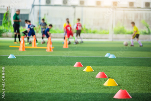 selective focus to red and yellow marker cones are soccer training equipment on green artificial turf with blurry kid players training background. material for training class of football academy.