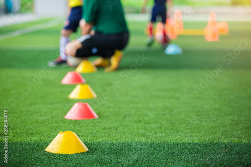 Selective focus to marker cones are soccer training equipment on green artificial turf with blurry coach is training kid players background. Material for training class of football academy. Vertical.