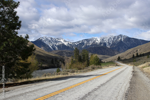 Adventure is out there: Beautiful landscape scenery of bare open highway road toward pine trees, green hills, and tall snow covered mountains in background under cloudy but sunny blue sky