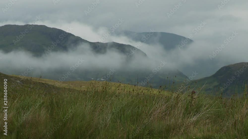 Steady, wide shot of grassy fields and thick fog in front of rolling mountains. Steady, wide shot of grassy fields and thick fog in front of rolling mountains.
