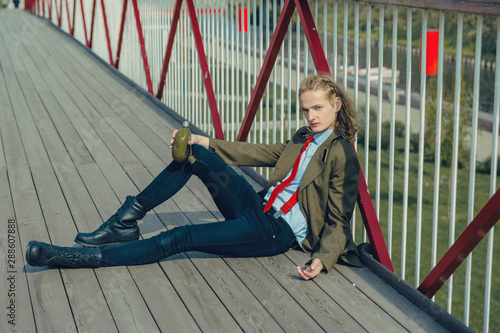 An attractive young man with long blonde hair, in berets and a military coat is leaning on a pyril bridge, in his hand is a green flask.  photo