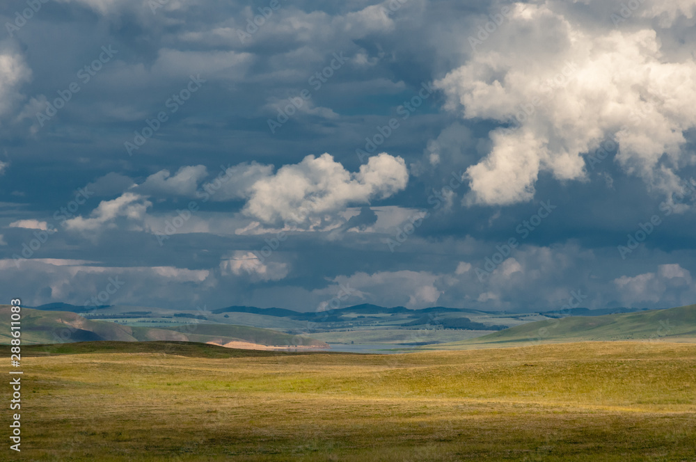 Panoramic landscape with steppe covered with green and yellow grass under blue sky with heavy clouds at Khakassia, Siberia, Russia.