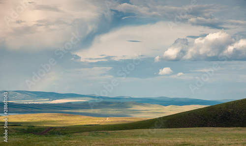 Panoramic landscape with steppe covered with green and yellow grass under blue sky with heavy clouds at Khakassia, Siberia, Russia.