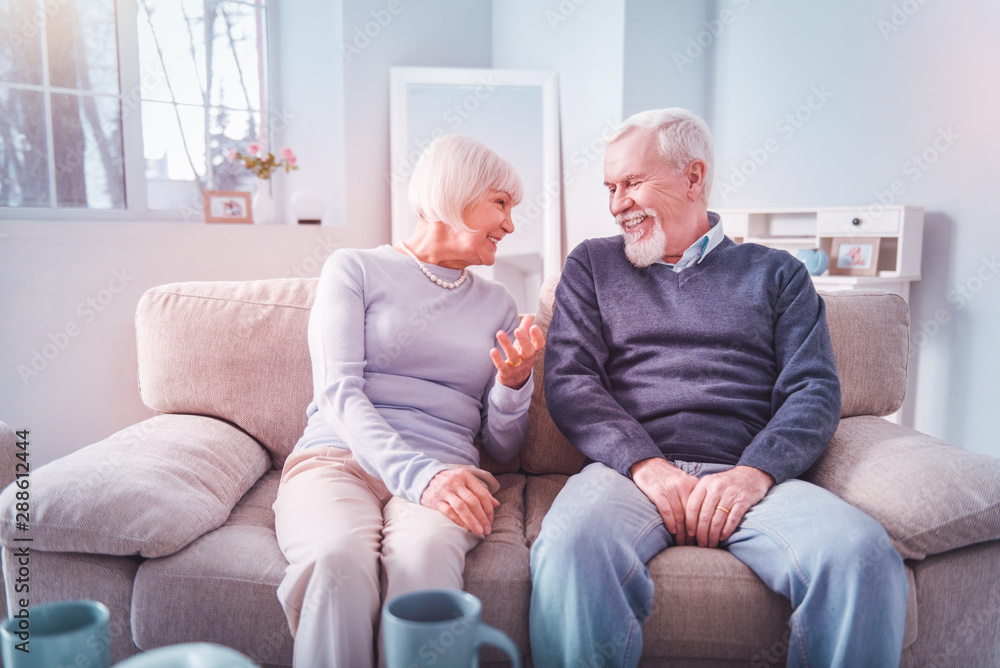 Couple of beaming retired man and woman sitting on sofa and chatting