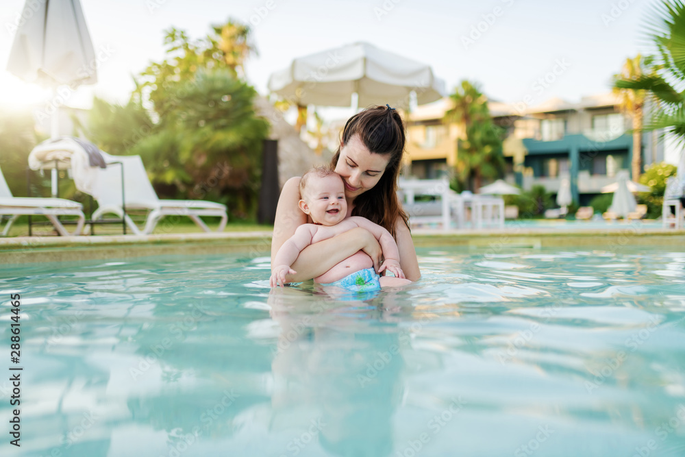 Beautiful Caucasian mother standing in swimming pool and holding her 6 months old son. Baby enjoying and smiling.