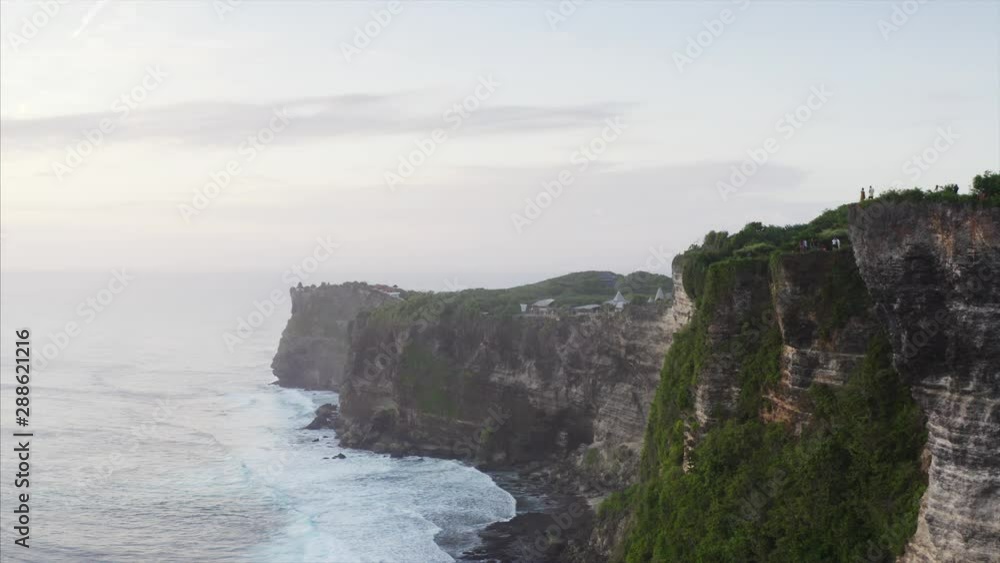 Aerial static view of high cliffs above blue ocean. Tourists on top of the cliffs are taking photos of landscape. Bali, Indonesia Aerial static view of high cliffs above blue ocean. Tourists on top of the cliffs are taking photos of landscape. Bali, Indonesia