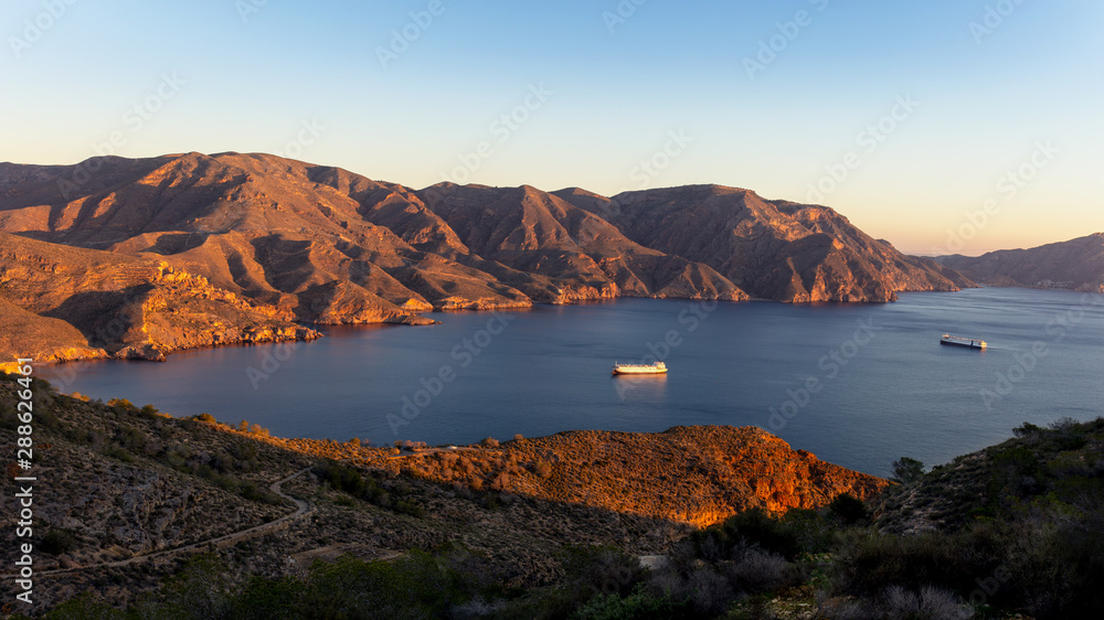 The morning sun shines into the bay at Cabo Tinoso near the Spanish port city of Cartagena. Large ships moored in the bay between the mountains.