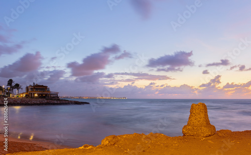 Morning just before sunrise on the Mediterranean south of the port city of Torrevieja. The still lit city can be seen in the background. In the foreground a small bay. Long Exposure.