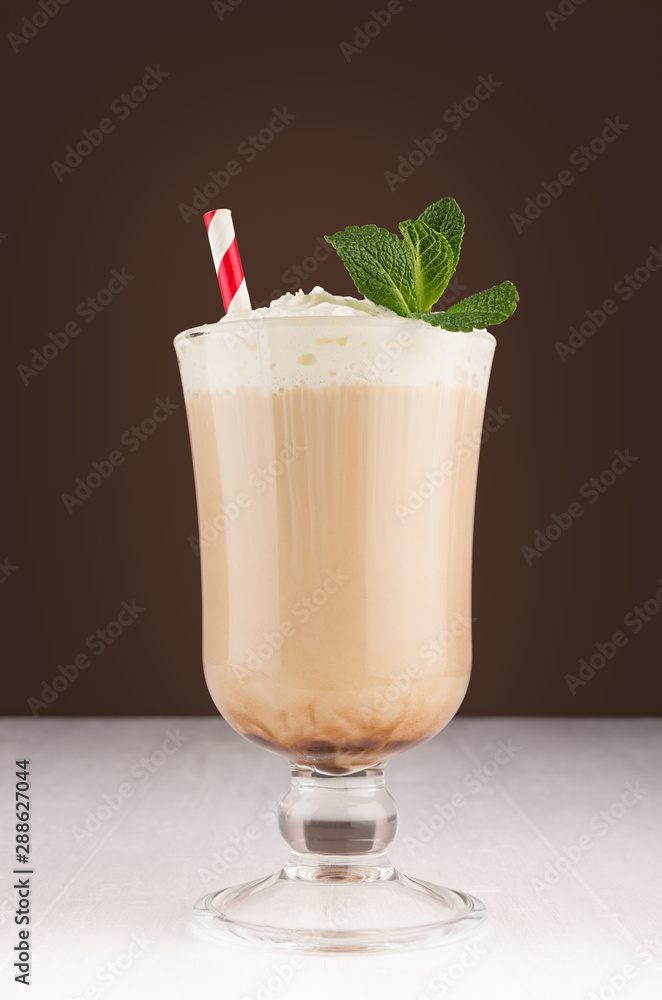 Macchiato coffee with layers, cream, red striped straw and green fresh mint in transparent goblet on dark brown background and white wooden table. Macchiato coffee with layers, cream, red striped straw and green fresh mint in transparent goblet on dark brown background and white wooden table.