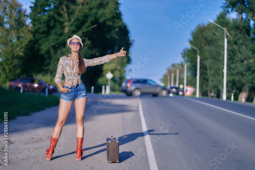 young beautiful girl stands on the road. plaid shirt  red boots and shorts. straw hat. travels. hitch-hiking. freedom. vacation.