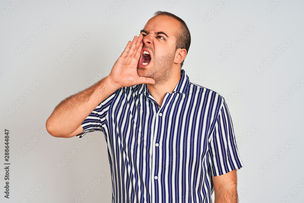 Young man wearing blue striped shirt standing over isolated white background shouting and screaming loud to side with hand on mouth. Communication concept. Young man wearing blue striped shirt standing over isolated white background shouting and screaming loud to side with hand on mouth. Communication concept.