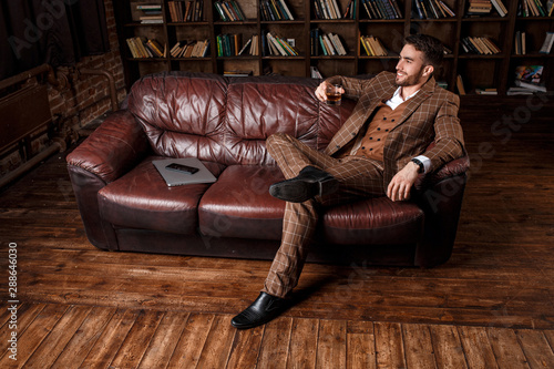 young successful man in a brown kosmtyum drinks whiskey. bearded businessman sitting in a library on a luxurious leather sofa and drinking cognac