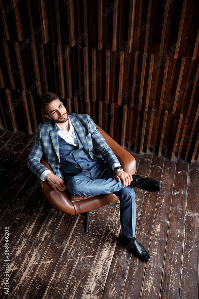 Young successful businessman in a blue suit in a cage is sitting in a leather chair. young man with a beard smiling. Young successful businessman in a blue suit in a cage is sitting in a leather chair. young man with a beard smiling.