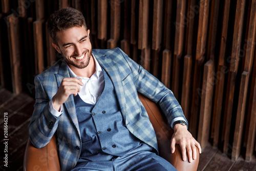 Young successful businessman in a blue suit in a cage is sitting in a leather chair. young man with a beard smiling and looks into the distance