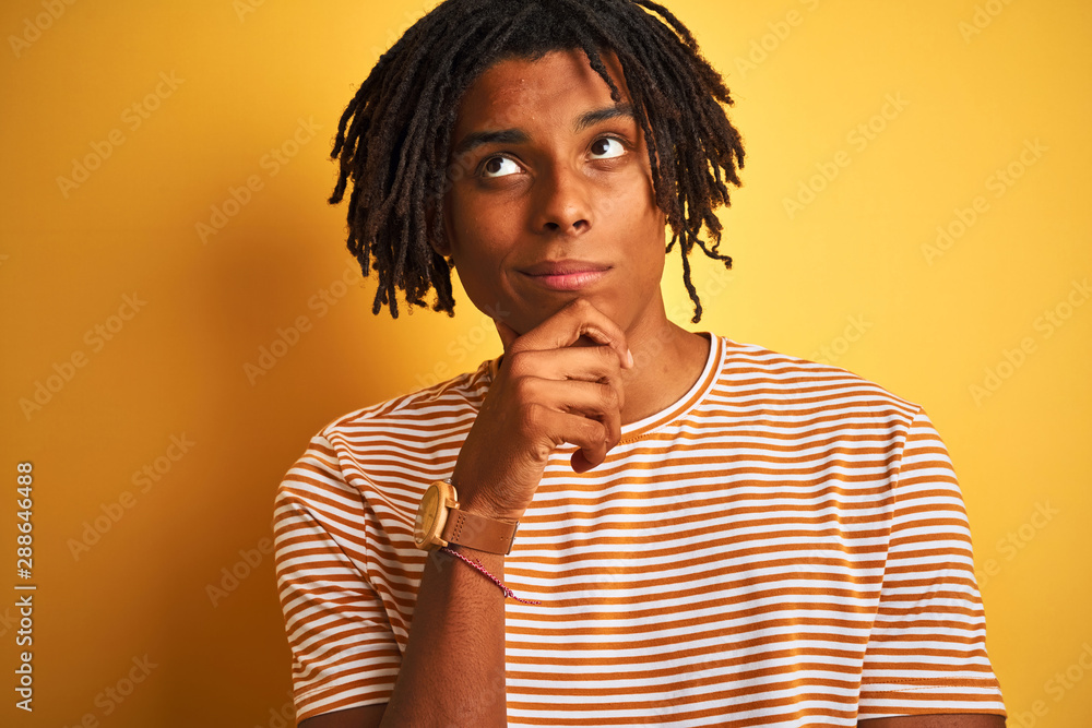 Afro man with dreadlocks wearing striped t-shirt standing over isolated yellow background serious face thinking about question, very confused idea Afro man with dreadlocks wearing striped t-shirt standing over isolated yellow background serious face thinking about question, very confused idea