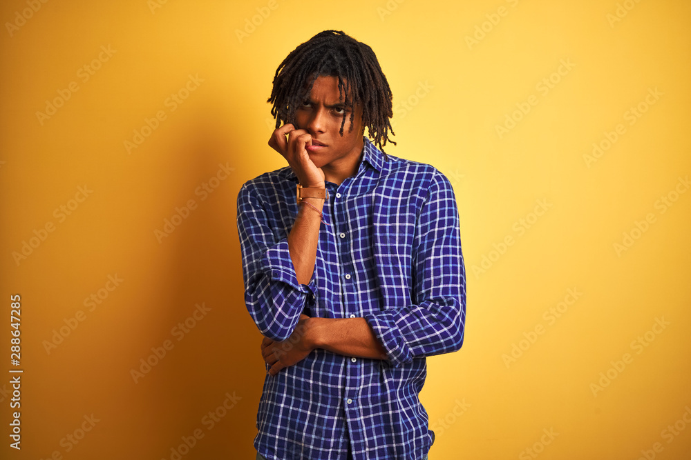 Afro man with dreadlocks wearing casual shirt standing over isolated yellow background looking stressed and nervous with hands on mouth biting nails. Anxiety problem. Afro man with dreadlocks wearing casual shirt standing over isolated yellow background looking stressed and nervous with hands on mouth biting nails. Anxiety problem.