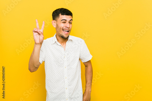 Young hispanic man winks an eye and holds an okay gesture with hand.